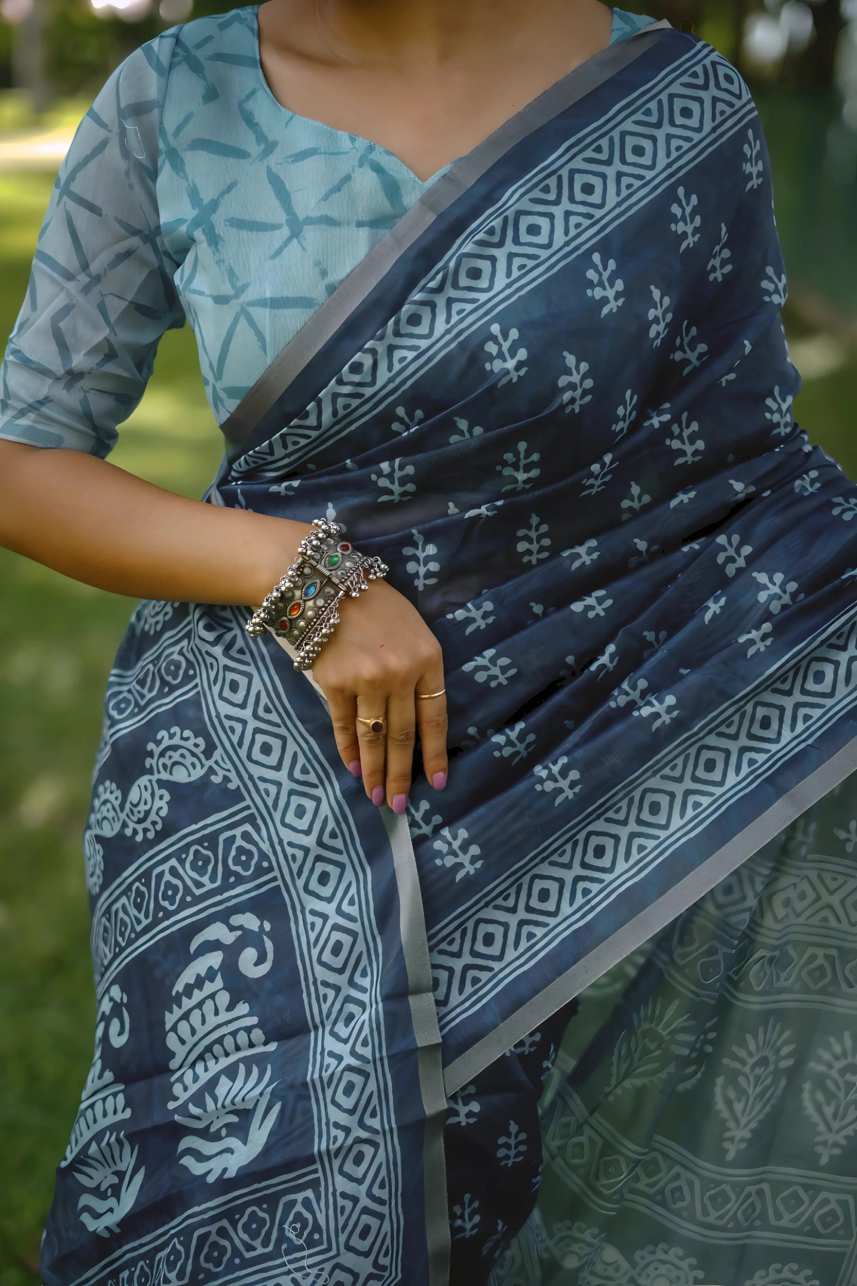 A woman models a striking Navy Blue Soft Cotton Saree featuring an all-over digital block print of traditional white motifs. She wears a light blue patterned blouse and silver jhumka earrings, posing on a path in a green outdoor setting.